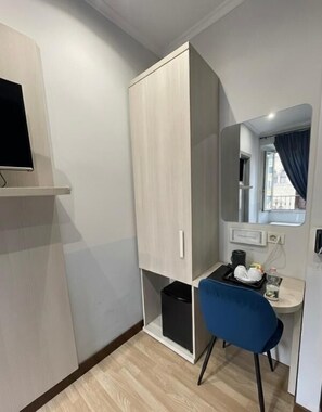 A compact room with a wardrobe, a small desk with a chair, a mirror, and a window with curtains.