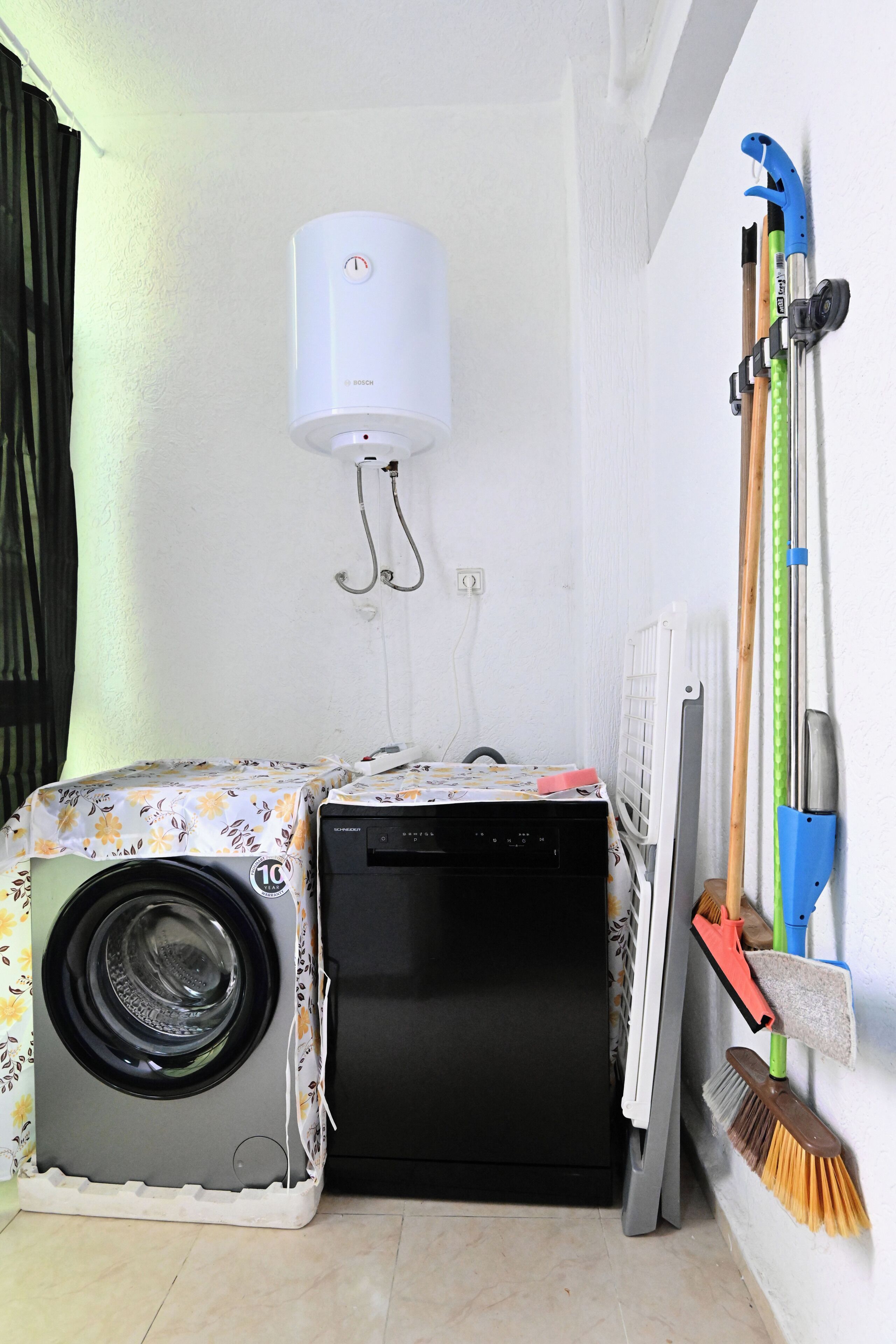 A compact laundry area with a Bosch washing machine and a dryer, a radiator, and cleaning tools on the wall.