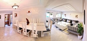 A spacious living area with a dining table and chairs, a sofa set, and a large window providing natural light.