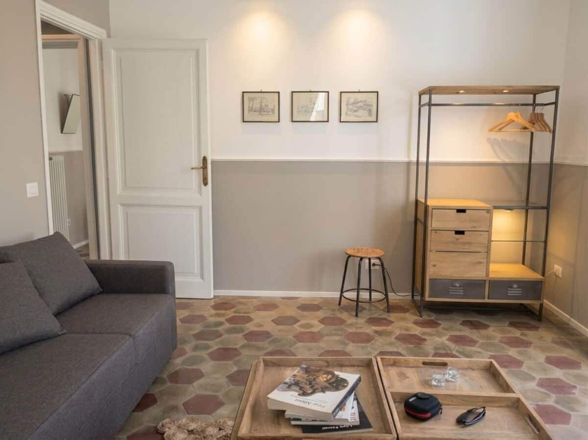 Un salon comprenant un canapé, une table basse avec des livres et une télécommande, un tabouret, un meuble de rangement en métal avec des vêtements suspendus et un sol à motifs.