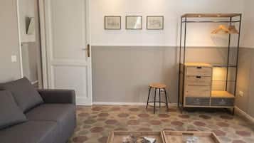 Un salon comprenant un canapé, une table basse avec des livres et une télécommande, un tabouret, un meuble de rangement en métal avec des vêtements suspendus et un sol à motifs.