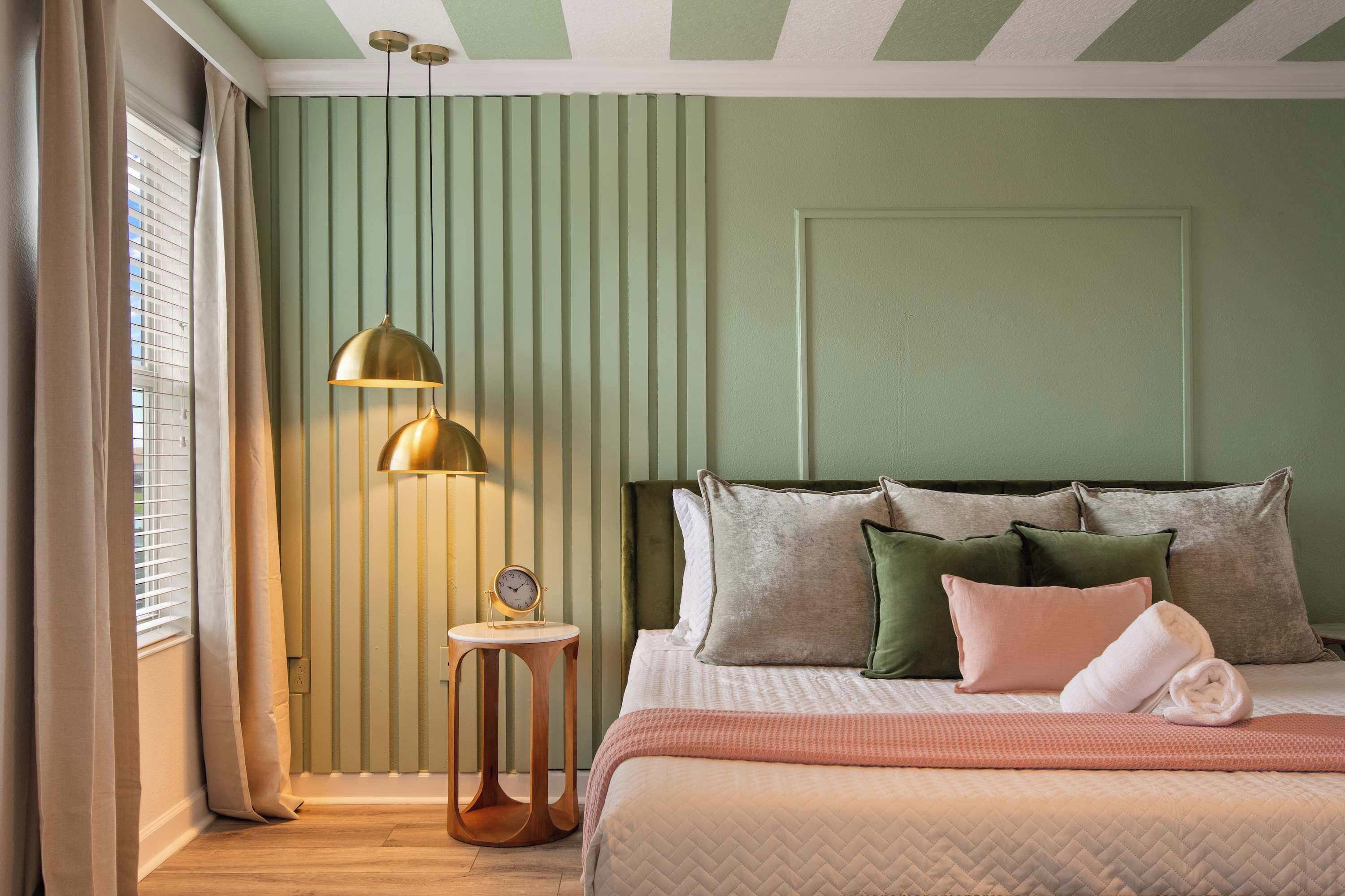 Un dormitorio con cama, almohadas, una mesita de noche con un reloj y una lámpara de pie.