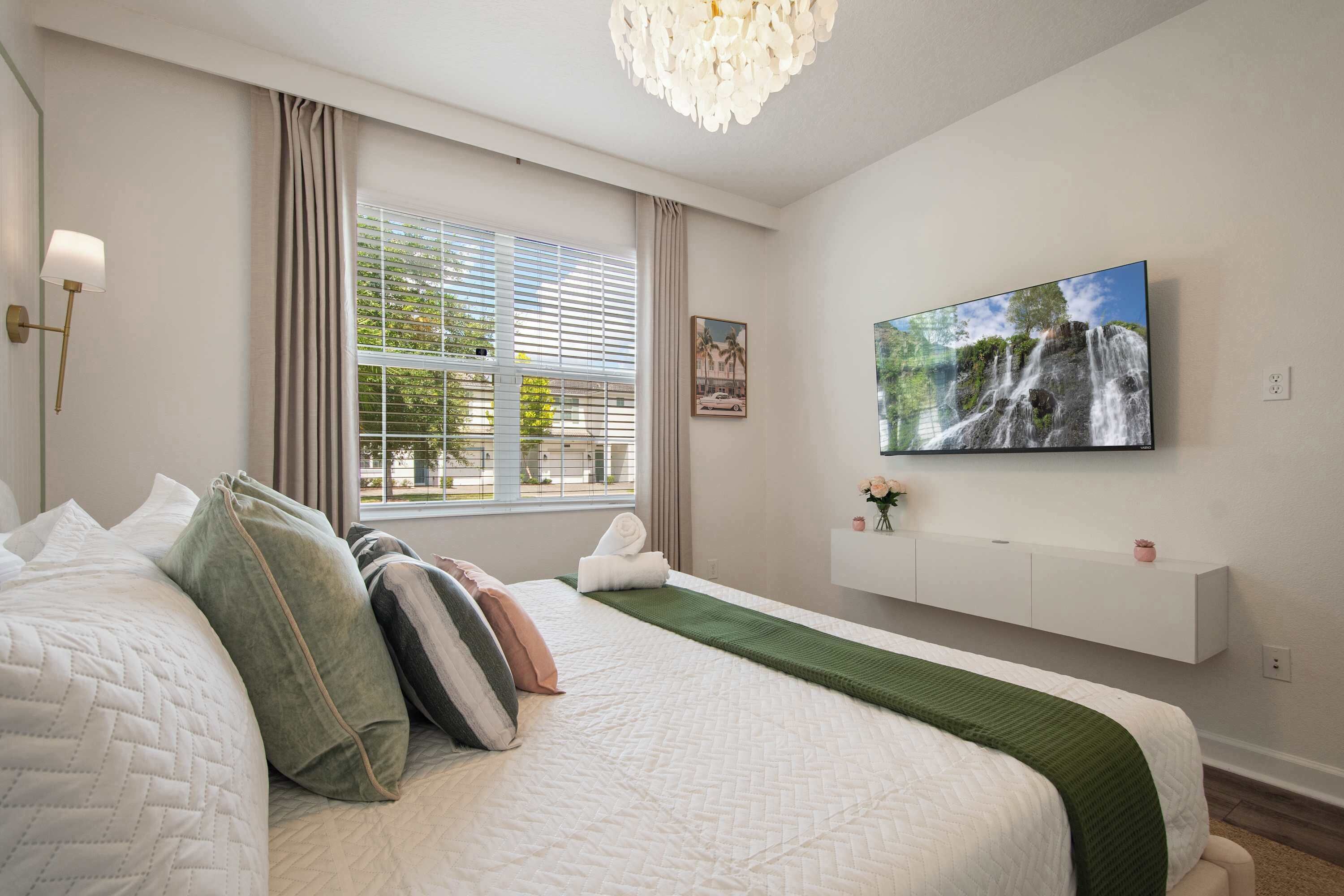Hotellihuone, jossa on suuri sänky, seinälle kiinnitetty televisio, ikkuna, jossa on sälekaihtimet, ja seinälle asennettu hylly, jonka päällä on maljakko.