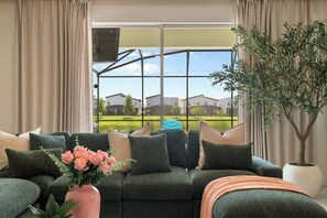 A living room with a sectional sofa, a potted plant, and a view of a residential area through large windows. - 6BR+Spa&Pool+Walk to Clubhouse+Theater+ThemedRooms (Kissimmee)