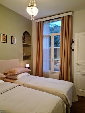 Ein Schlafzimmer mit zwei Betten, einer Kristallleuchter, einem Fenster mit Vorhängen und einer Tür mit Kleiderhaken.