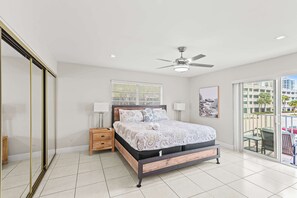 Ein Schlafzimmer mit einem Bett, Nachttischen, einem Deckenventilator, einem Fenster mit Jalousien und Blick ins Freie.