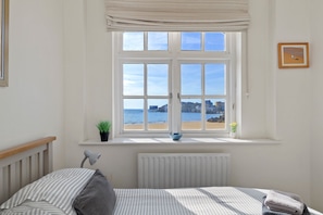 Een slaapkamer met een groot raam, een bed, een radiator, een plant in pot en een ingelijst kunstwerk aan de muur.