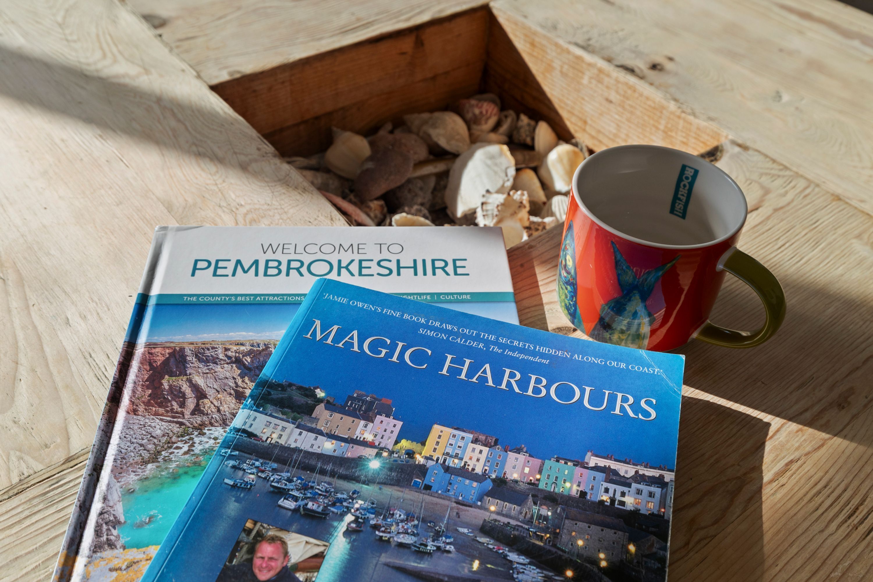 Two travel books titled 'Welcome to Pembrokeshire' and 'Magic Harbours' on a wooden surface with a red mug and seashells.