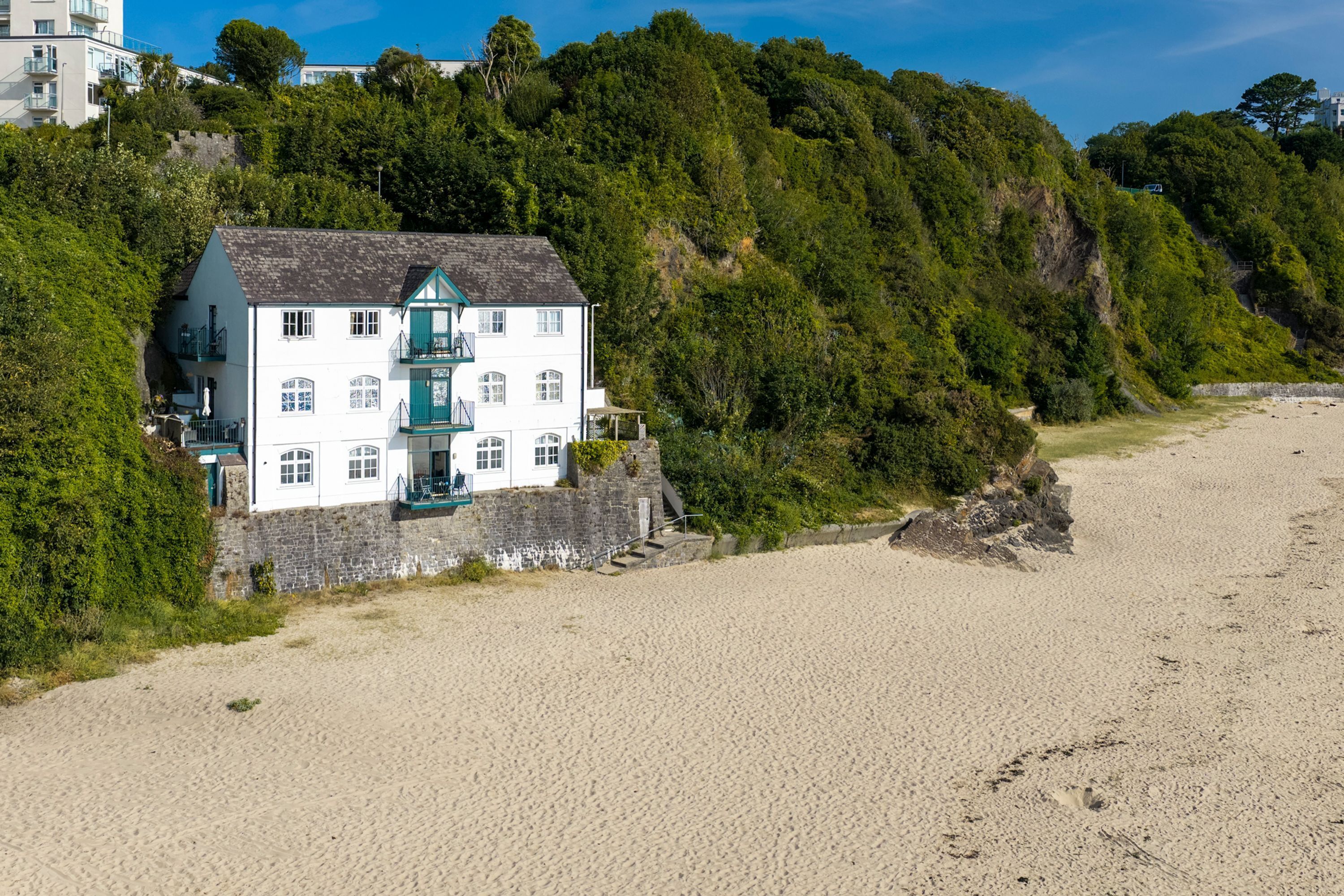 Ein weißes Gebäude mit einem grünen Balkon, gelegen an einem Sandstrand mit einem bewaldeten Hügel im Hintergrund.
