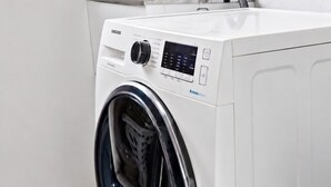A front-loading washing machine in a laundry room with a tiled wall and a window. - Bright 1-bed Retreat in Kingsville (Melbourne)