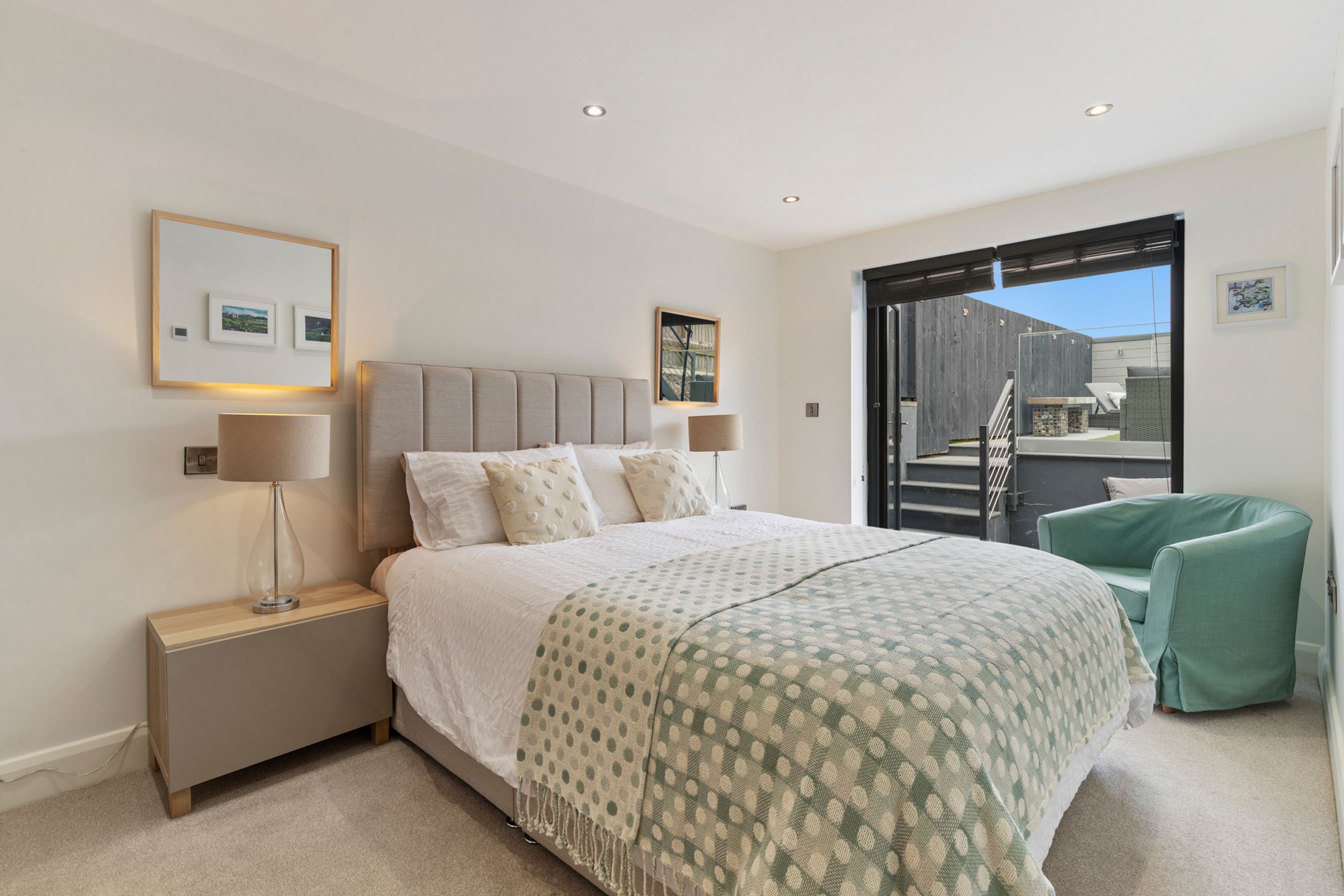 Une chambre à coucher avec un lit, des lampes de chevet, une table de chevet, un fauteuil vert et une vue sur l’extérieur.