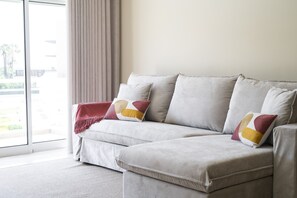 A neutral-toned sofa with a throw blanket and decorative pillows, positioned in a room with a large window and a view of greenery outside. - Moura Praia by Hdproperties (Vilamoura)