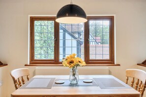 A dining table with a vase of sunflowers, two chairs, a pendant light, and a view of a building through two windows. - 2-bed - Sleeps 4 - Parking & Ev Charging (Wadebridge)