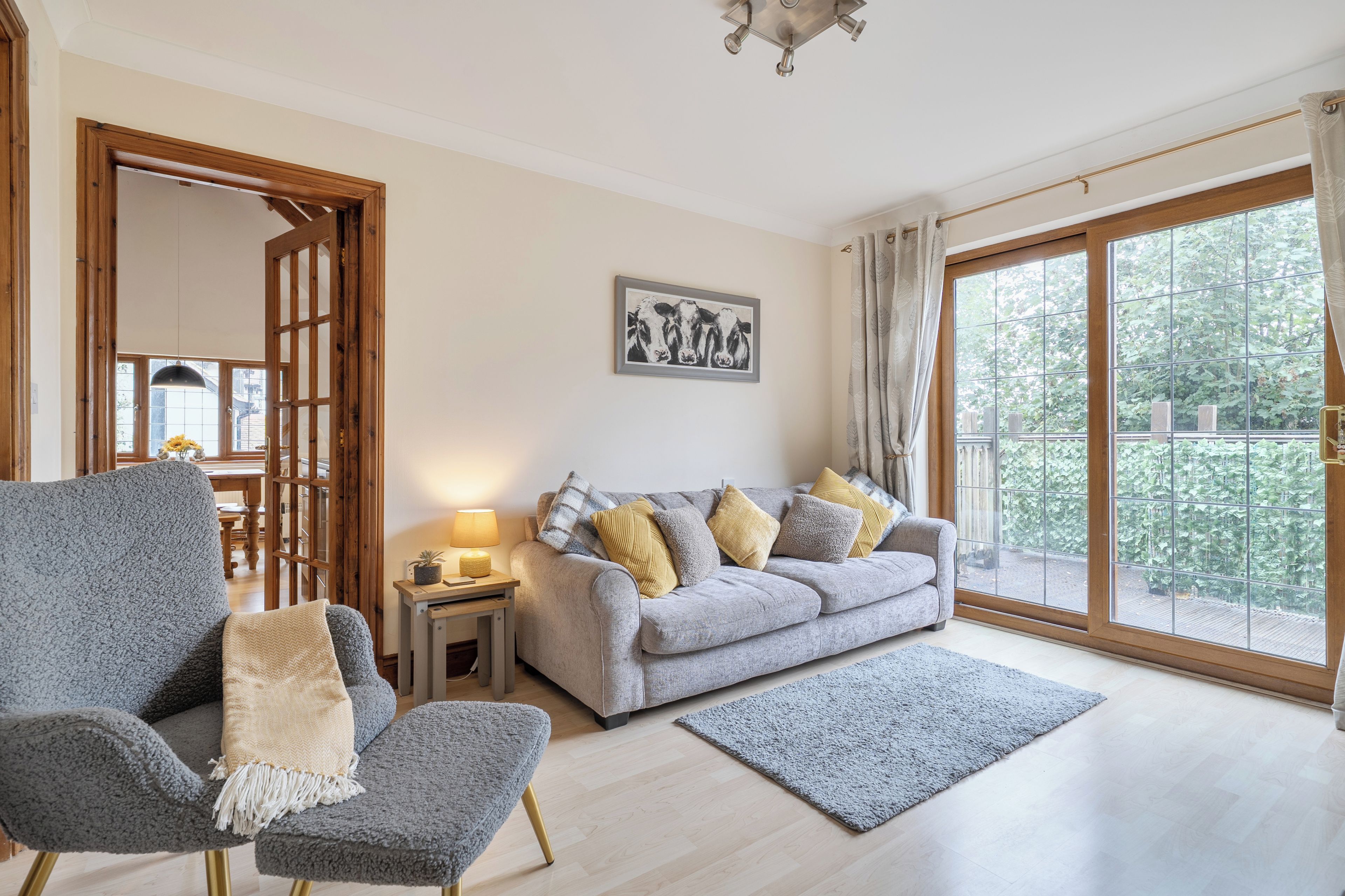 Uma sala de estar com sofá cinza, poltrona cinza, mesinha lateral com abajur, quadro pendurado na parede e uma grande porta de vidro deslizante para o exterior.