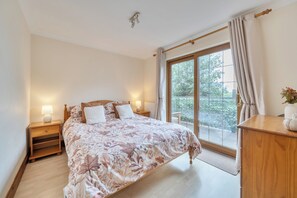 A bedroom with a large bed, a nightstand with a lamp, a wooden dresser, and a view of greenery through a sliding glass door. - 2-bed - Sleeps 4 - Parking & Ev Charging (Wadebridge)