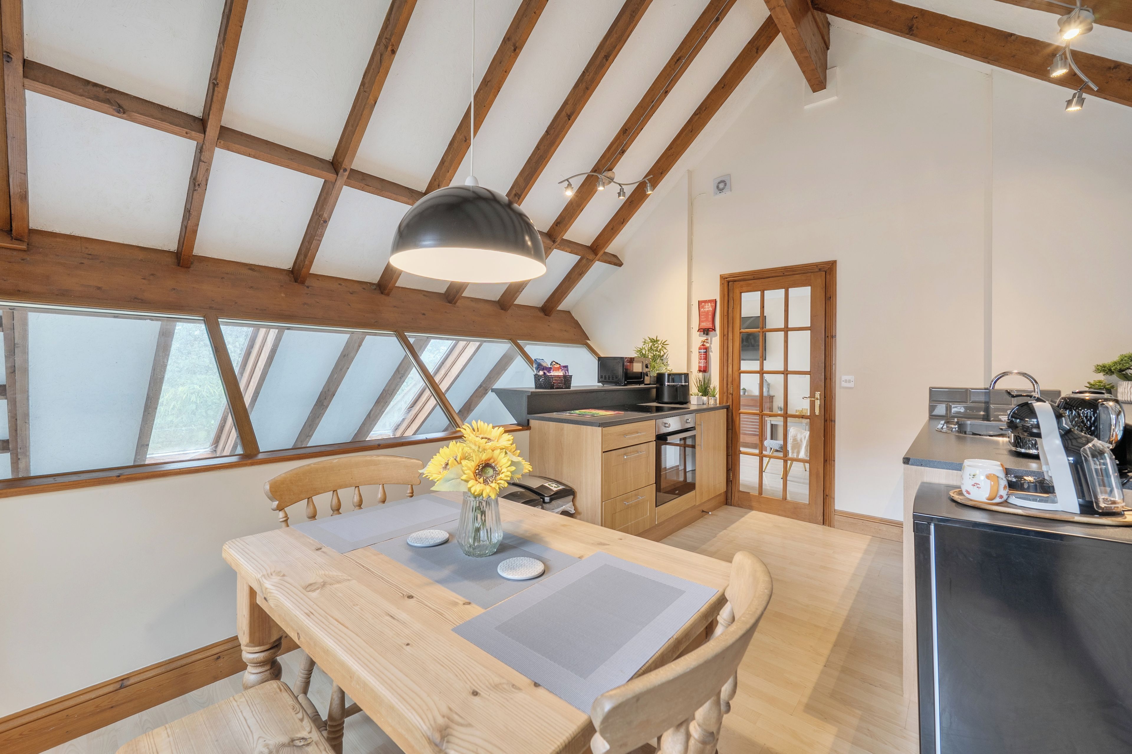 Una cocina con vigas de madera, una mesa de comedor con sillas, un florero con flores y vistas a través de las ventanas.