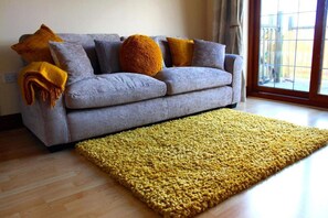 A living room with a grey sofa, a yellow rug, and a window with a view. - 2-bed - Sleeps 4 - Free Parking & Ev Charging (Wadebridge)
