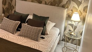 A bedroom with a bed, pillows, a bedside table with a lamp, and a wall with a leaf pattern. - Le Petit Rocher Doré: a unique getaway with a view of the Rock. (Pierrelatte)