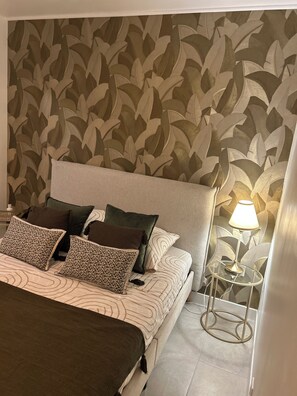 Ein Schlafzimmer mit einem Bett, Kissen, einem Nachttisch mit Lampe und einer Wand mit Blättermotiv.
