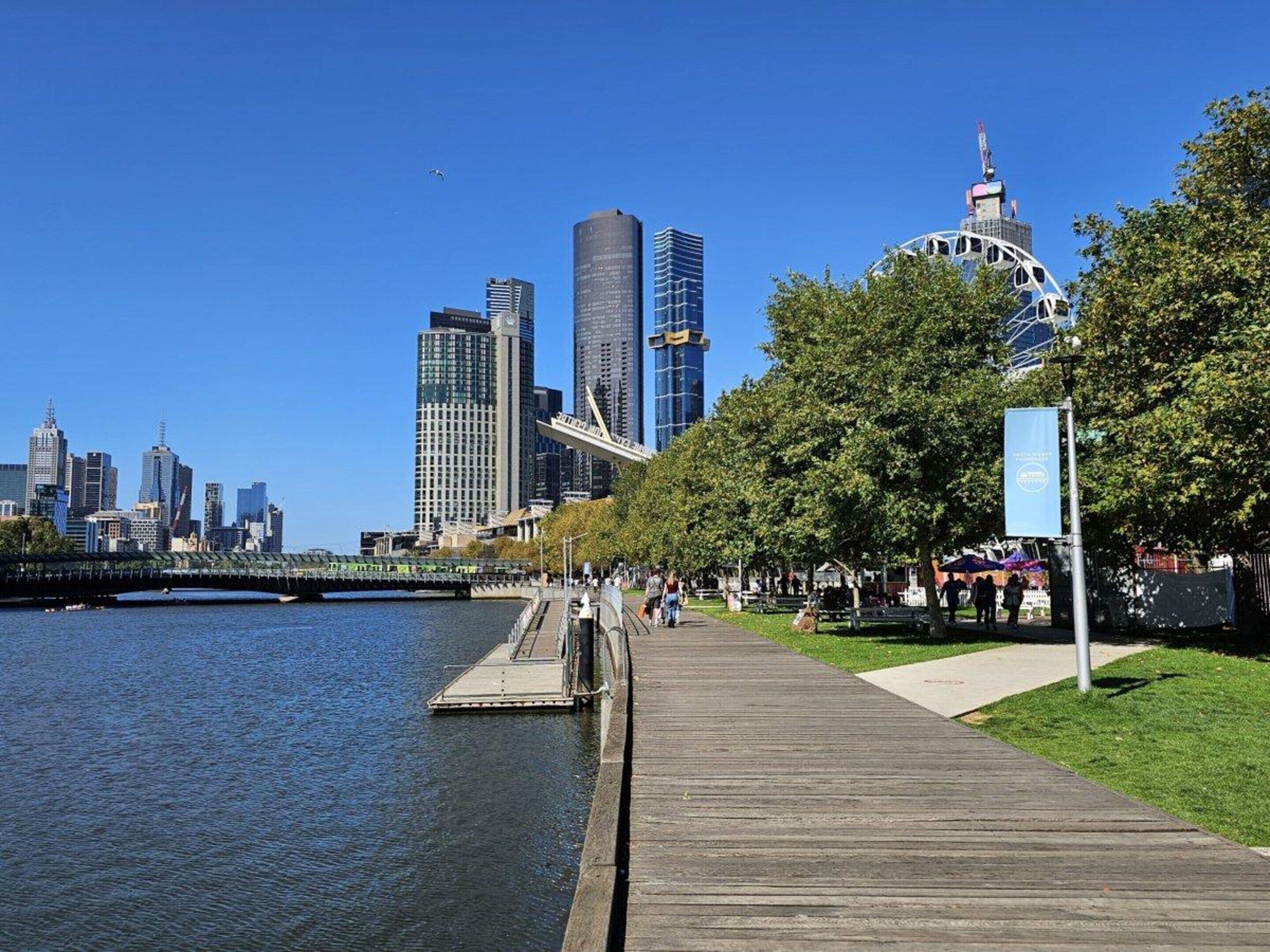 A waterfront promenade with a wooden boardwalk, a river, and a city skyline in the background.