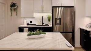 A modern kitchen with white cabinetry, a stainless steel refrigerator, a marble countertop, and a hanging plant. - Luxury 2BR • 2.5 Bath Designer Home Retreat (Montréal)