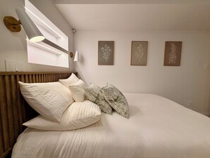 A neatly made bed with white bedding and decorative pillows, a wall-mounted light fixture, and three framed botanical prints on the wall. - Luxury 2BR • 2.5 Bath Designer Home Retreat (Montréal)