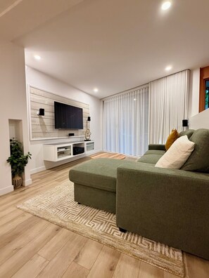 A modern living room with a large flat-screen TV mounted on a white wall, a green sofa with cushions, and a patterned rug on the wooden floor. - Luxury 2BR • 2.5 Bath Designer Home Retreat (Montréal)