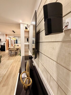 A modern living room with a flat-screen TV mounted on a textured wall, a dining area with a table and chairs, and a kitchen in the background. - Luxury 2BR • 2.5 Bath Designer Home Retreat (Montréal)