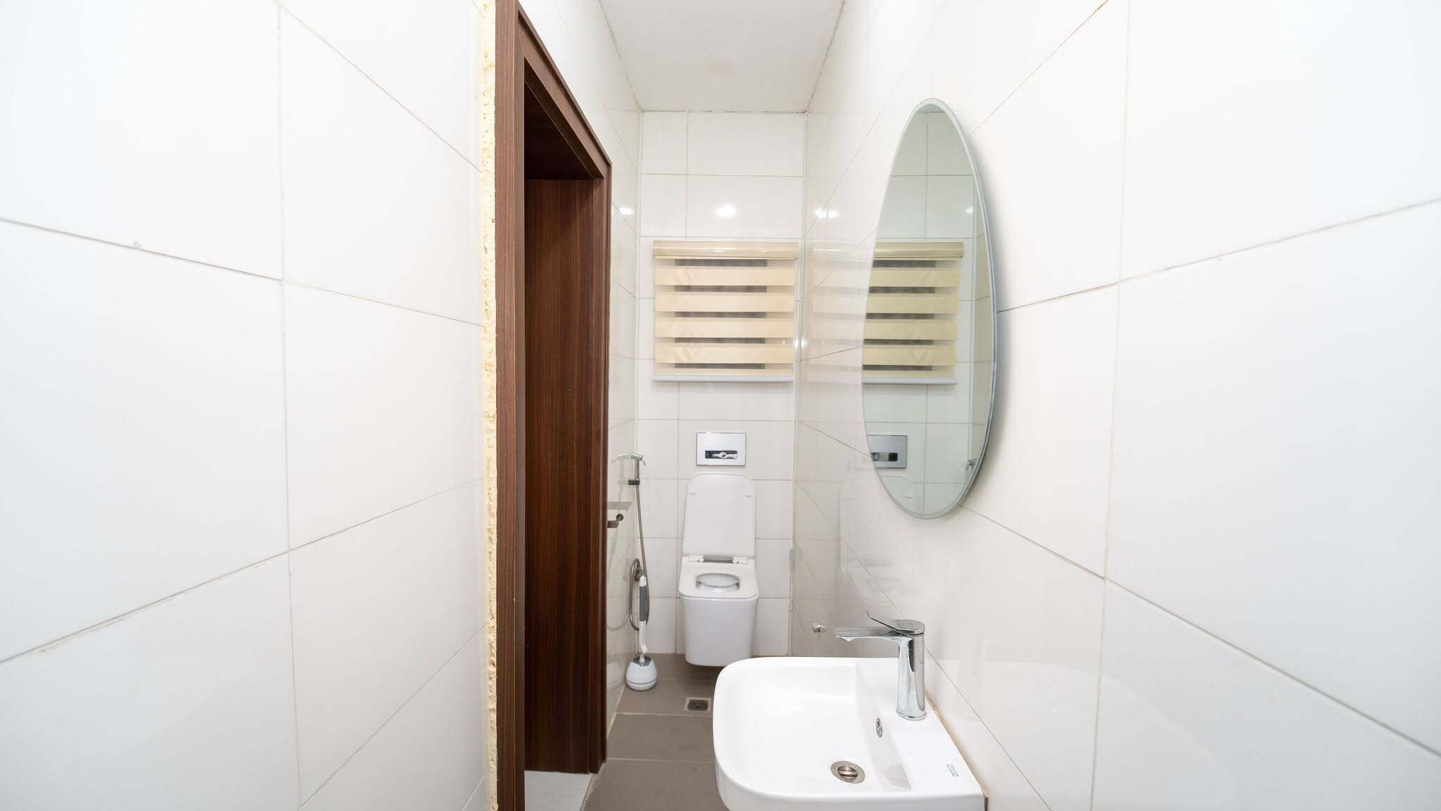A bathroom with white tiled walls, a wooden door, a white toilet, a bidet, a mirror, and a sink.