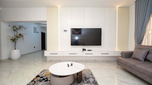Una sala de estar moderna con un televisor de pantalla plana montado en una pared blanca, una mesa redonda de centro, un sofá y una planta en maceta.