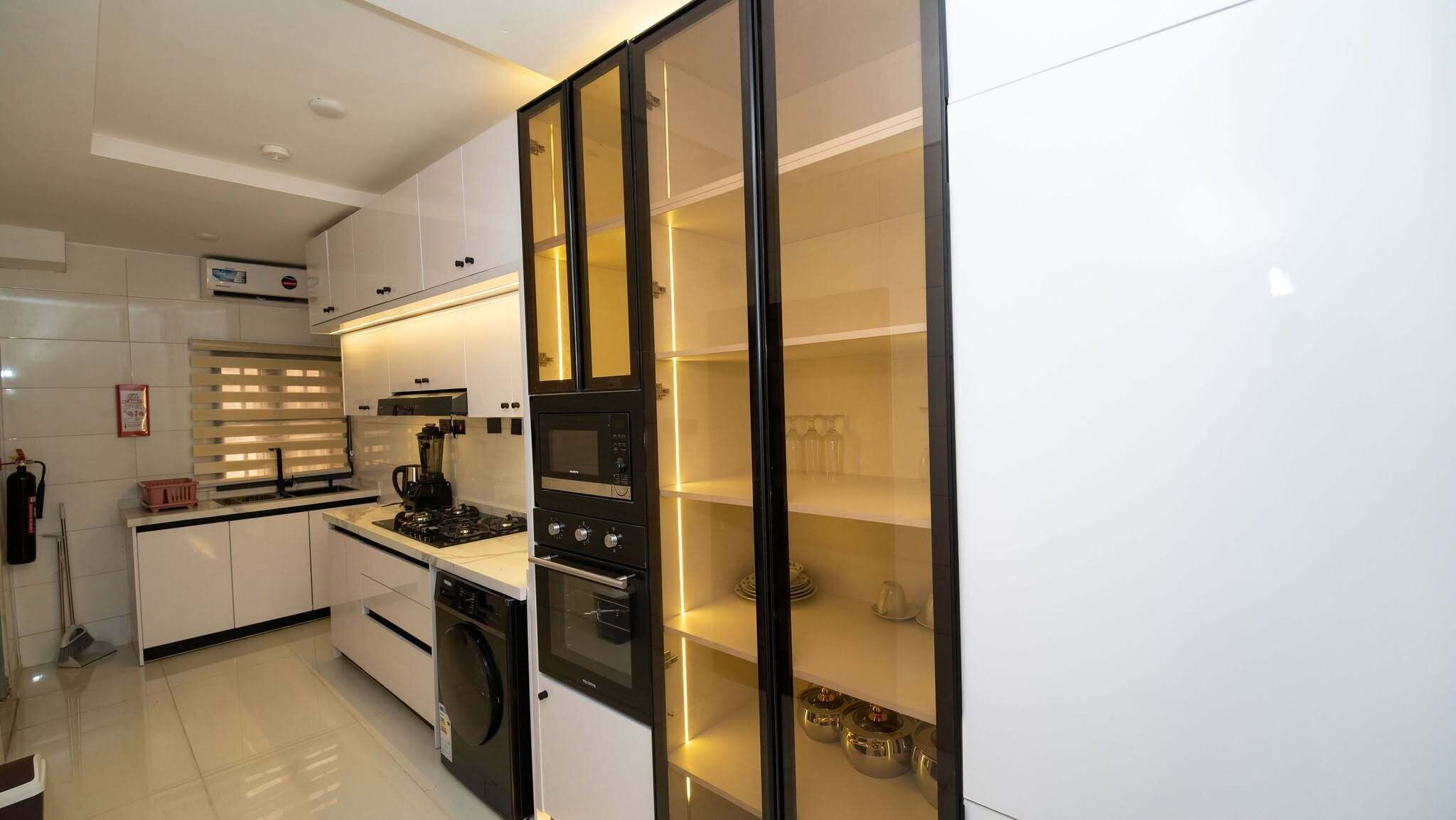 Una cucina moderna con mobili bianchi, forno integrato, macchina per il caffè e pensile in vetro con stoviglie.