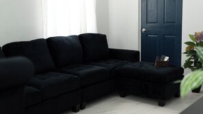 A black sofa and ottoman in a room with a blue door and a basket with items on the floor. - Cozy 1-bedroom house in charming Soufrière with AC (Soufrière)