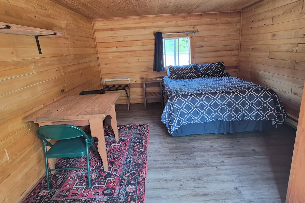 Una habitación de cabaña de madera pequeña con una cama, un escritorio, una silla y un ventanal.