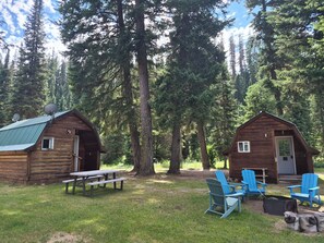 Two wooden cabins with satellite dishes, a picnic table, and blue Adirondack chairs in a forest setting.