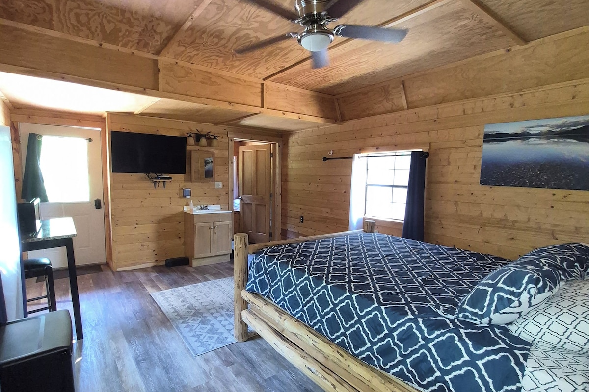 Interior de una cabaña de madera con cama, escritorio y ventilador de techo.