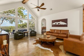 A spacious living room with a large window, leather furniture, a ceiling fan, and a cow silhouette artwork. - Cliffside Lodge + Hill Country Views (Wimberley)