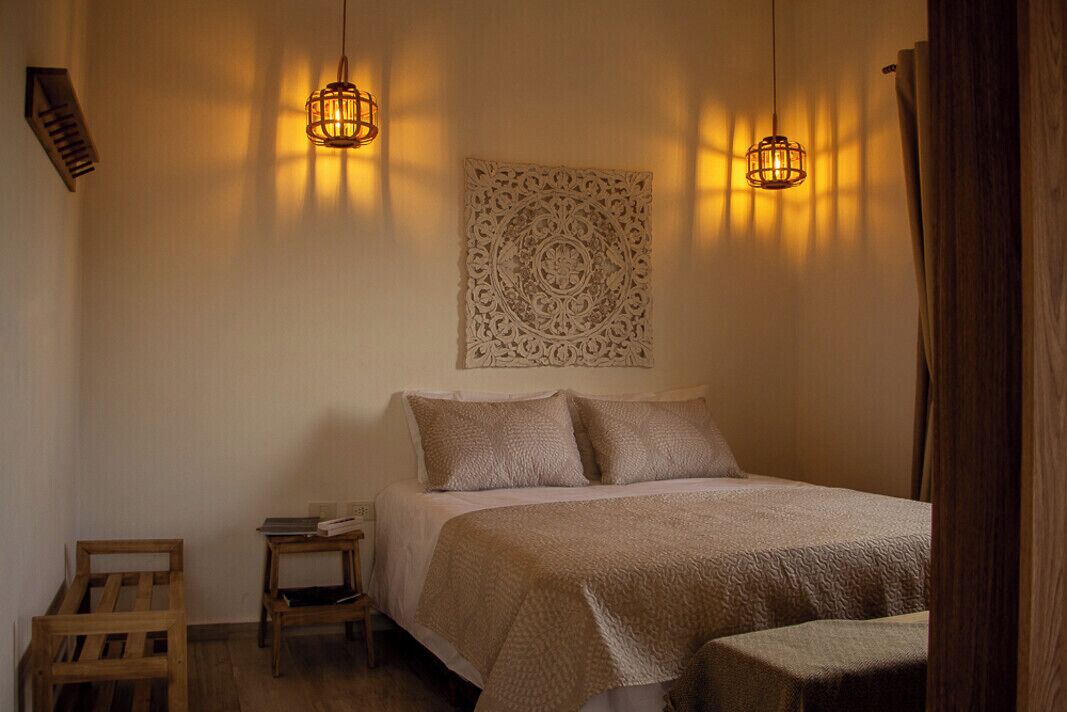 Um quarto com cama, duas mesas de cabeceira, um objeto decorativo na parede e dois lustres.