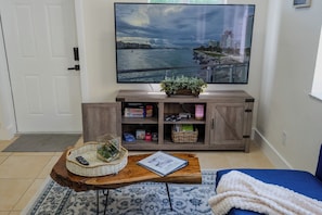 A living room with a television mounted on the wall, a wooden coffee table with a basket and a book, a blue sofa, and a cabinet with books and decorative items. - Gateway to the Keys - Private Family Retreat (Homestead)