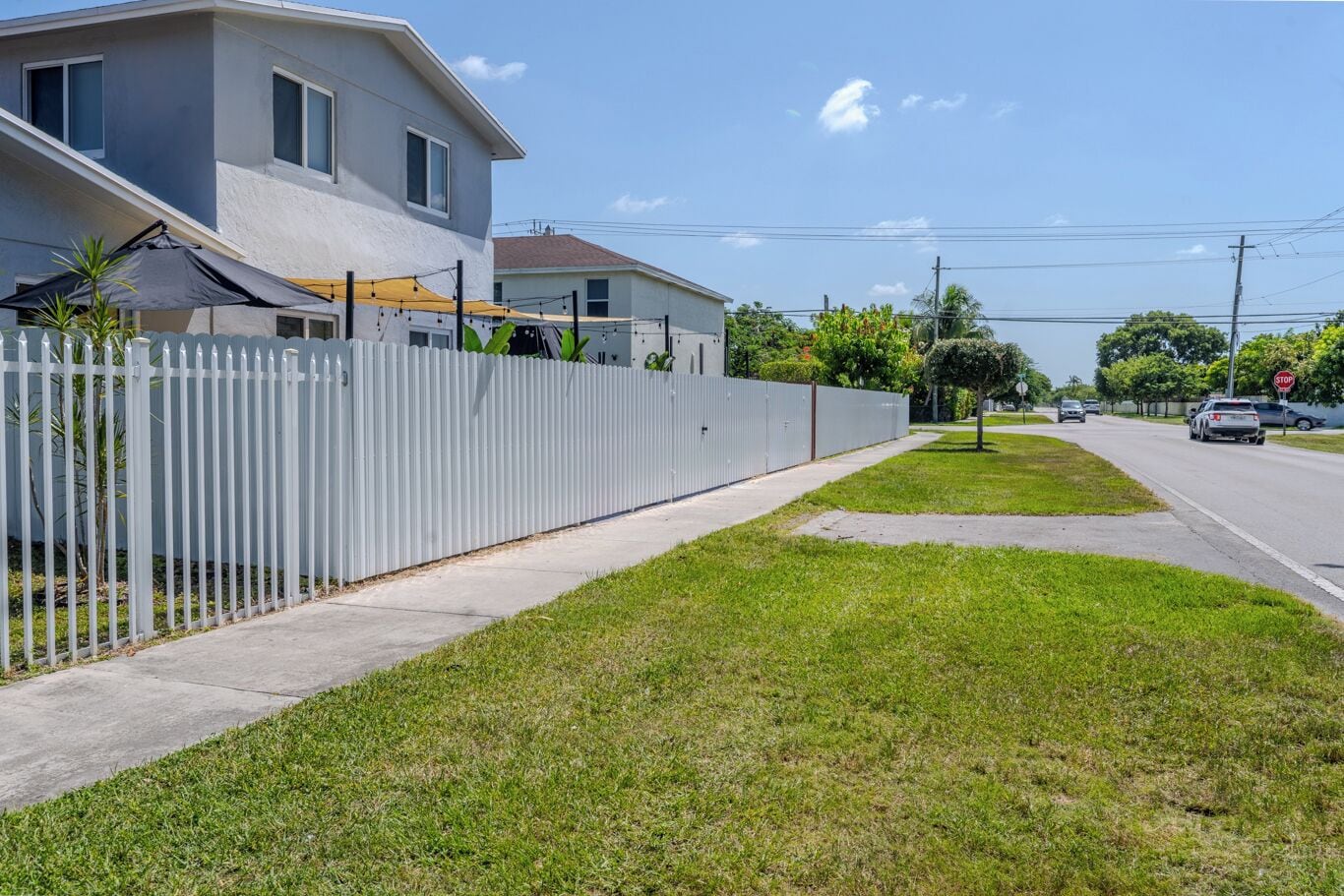 A residential street with a white fence, a sidewalk, and houses with visible windows and a covered patio.
