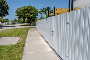A concrete sidewalk adjacent to a corrugated metal fence, with a grassy area and trees in the background. - Gateway to the Keys - Private Family Retreat (Homestead)
