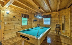 A wooden room with a pool table, two windows with curtains, and a hanging light fixture.