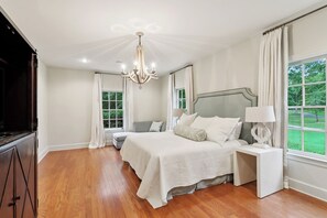 A bedroom with a large bed, bedside tables, a chandelier, and a view of greenery through the window. - Spacious 7BR Country Retreat w/ Pool, Tennis Court & Private Pond in Folsom, LA (Folsom)