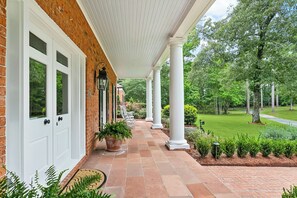 Eine überdachte Veranda mit weißen Säulen, eine Backsteinmauer und ein Garten mit Rasen und Bäumen.