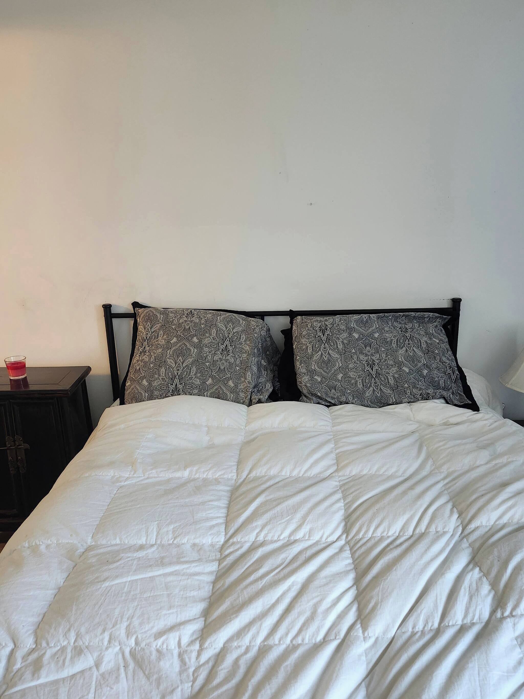 A bed with a white quilted bedspread and two patterned pillows, a dark wooden nightstand with a red object on it, and a white wall.