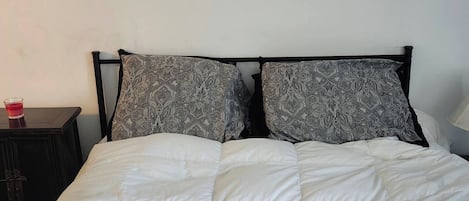 Una cama con una colcha blanca acolchada y dos almohadas estampadas, una mesita de noche de madera oscura con un objeto rojo encima y una pared blanca.