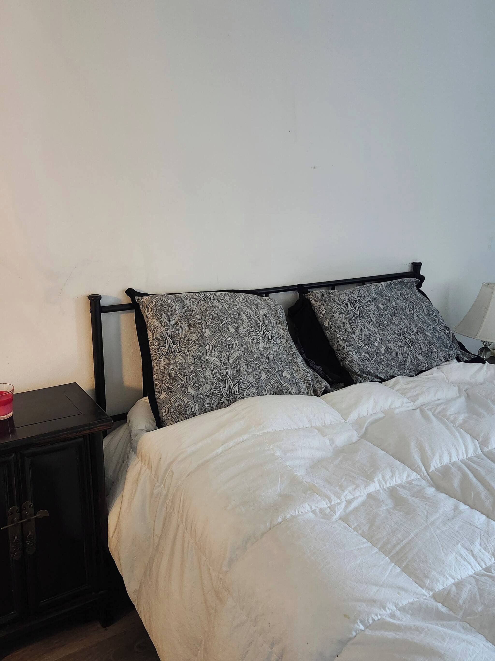Una cama con edredón blanco y dos almohadas estampadas, una mesita de noche pequeña con un objeto rojo, y un mueble auxiliar de madera oscura.