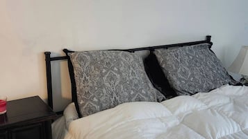 Una cama con edredón blanco y dos almohadas estampadas, una mesita de noche pequeña con un objeto rojo, y un mueble auxiliar de madera oscura.