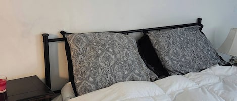 Una cama con edredón blanco y dos almohadas estampadas, una mesita de noche pequeña con un objeto rojo, y un mueble auxiliar de madera oscura.