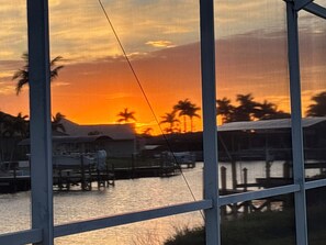 A sunset view through a window, with a dock and boats in the foreground and palm trees in the background. - Sun-Soaked SW Cape Coral Gulf Access Retreat (Cape Coral)