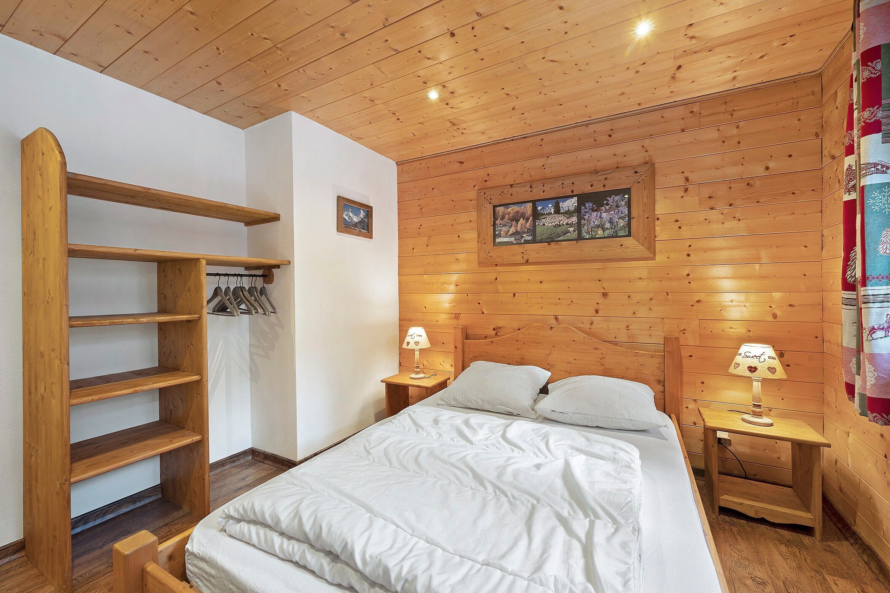 A wooden-paneled room with a bed, two bedside tables, a wooden shelf unit, and a framed picture on the wall.